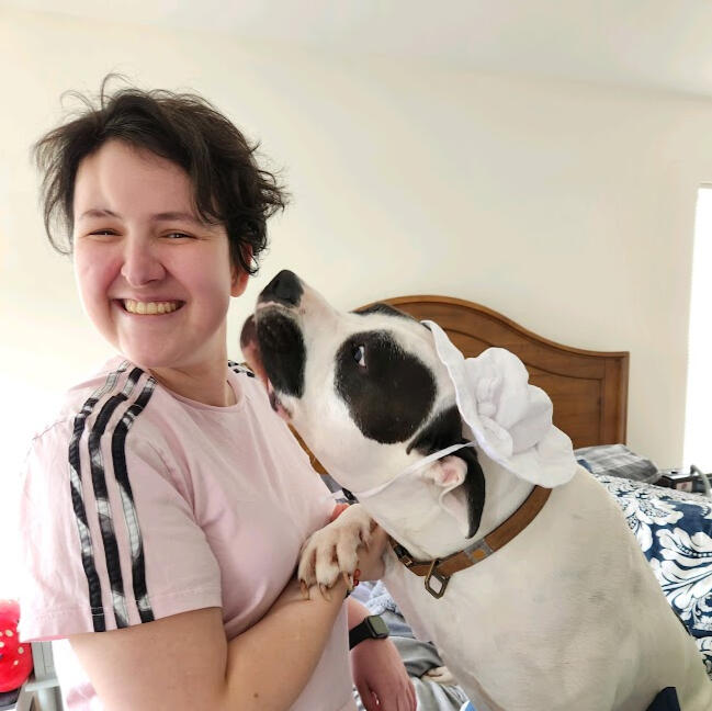 an image of Mack in a pink shirt with black stripes down the sleeve turned sideways smiling at the camera. Their dog is on the bed in front of them, paw in their hand, wearing a toddler's white hat, trying to lick their face.